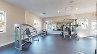 This single-level fitness center is designed to support a comprehensive workout experience, combining functional layout with a bright, motivating atmosphere. The space is finished with durable dark rubber flooring throughout, providing a practical and resilient surface suited for heavy equipment and high-traffic use.

The strength training area is well-appointed with a variety of machines, including a cable crossover/Smith machine combo, a lat pulldown station, a seated press machine, and a weight bench with plate-loaded barbells. A large wall-spanning mirror runs along one side of the room, visually expanding the space and allowing users to monitor their form during exercises.

The cardio zone features multiple treadmills aligned along a mirrored wall, alongside additional cable resistance machines. Large windows and glass door panels allow generous natural light to fill the room while offering views of the surrounding outdoor environment.

A standout design element is a bold geometric accent wall painted in vivid yellow and navy blue, adding visual energy and a modern aesthetic to the space. Recessed lighting and ceiling-mounted ventilation ensure the environment remains well-lit and comfortable. The overall ambiance is clean, open, and purpose-driven, making this fitness center a fully functional facility for both cardiovascular and strength training workouts.