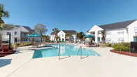 Located at 650 W Flintlake Ct in Myrtle Beach, South Carolina, this outdoor community amenity area centers around a generously sized, freeform resort-style swimming pool with crystal-clear blue water. The expansive concrete pool deck provides ample room for lounging, with multiple teal-umbrella shaded seating areas and reclining lounge chairs arranged throughout, offering both sun and shade options for residents.

The pool area is enclosed by a white metal fence, maintaining a sense of privacy and safety while preserving open views of the surrounding residential buildings and well-maintained landscaping. Towering palm trees and manicured shrubs accent the deck, lending a distinctly coastal, tropical ambiance that complements the Myrtle Beach setting.

Adjacent to the pool is a covered pavilion attached to the community clubhouse, supported by classic white columns. This shaded area features high-top seating, a water fountain, posted pool rules, and convenient vending. A set of elegant green French doors provides access to the clubhouse interior, extending the space's functionality beyond the pool itself.

Under the clear South Carolina sky, the entire area exudes a bright, open, and inviting atmosphere. The combination of lush palm trees, well-kept grounds, and the multi-building residential backdrop creates a cohesive and appealing outdoor retreat for the community.