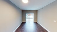 This single-floor unit offers a well-designed layout that balances functionality and comfort across its living, sleeping, kitchen, and bathroom spaces. The main living area is finished with rich, dark hardwood-style flooring that extends throughout much of the unit, complemented by neutral taupe and gray wall tones. A sliding glass door fitted with sheer vertical blinds floods the living space with natural light and provides direct views of the surrounding neighborhood.

The kitchen features a practical galley-style configuration with dark espresso cabinetry, granite-patterned countertops, a stainless steel double sink, and a full suite of stainless steel appliances including a refrigerator, range, and microwave. An in-unit laundry area with a washer and dryer is conveniently accessible directly from the kitchen. A peninsula counter provides additional prep space and separates the kitchen from the adjacent living area.

The bedroom is finished with plush wall-to-wall carpeting and features a window with horizontal blinds, allowing natural light while maintaining privacy. The bathroom includes a spacious walk-in shower enclosed by sliding glass doors with chrome hardware, white tiled walls, and built-in shelving, alongside safety grab bars for added accessibility.