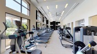 This contemporary fitness center is designed to deliver a premium workout experience across a single, thoughtfully organized floor. The cardio area is a standout feature, showcasing a long row of treadmills, ellipticals, and stationary bikes neatly arranged along a wall of expansive floor-to-ceiling windows. These large windows flood the space with natural light and offer views of lush outdoor greenery, creating an energizing atmosphere for exercise. Overhead, recessed panel lighting and a large ceiling fan provide additional comfort and ventilation throughout the cardio zone. The light wood-toned flooring runs throughout, lending a clean, modern aesthetic that complements the neutral cream and black color palette. Adjacent to the cardio area, a dedicated strength and functional training zone features multi-rack systems, resistance equipment, barbells, plyo boxes, and stretching mats. This area benefits from impressively high ceilings with clerestory windows, amplifying the sense of openness. Mirrored walls further enhance the spaciousness while providing visual feedback for proper form. The overall ambiance is bright, airy, and professional, making this fitness center a well-rounded facility suited for a wide range of training styles and fitness levels.