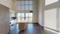 Located on the iconic Wilshire Boulevard in Los Angeles, this modern two-story residence showcases a thoughtfully designed open layout with impressive double-height ceilings that create a dramatic sense of volume in the main living area. Expansive floor-to-ceiling windows and sliding glass doors flood the space with natural light and open onto a balcony offering sweeping city views, making the living and dining area a true centerpiece of the home. Rich dark hardwood-style flooring flows throughout, complementing the neutral warm-toned walls. A freestanding wood-and-white island on casters adds versatile workspace to the open-plan area. The kitchen is well-appointed with glossy upper cabinetry, warm wood lower cabinets, a multi-toned glass tile backsplash, white quartz countertops, and a full suite of stainless steel appliances including a gas range, built-in microwave, refrigerator, and dishwasher. The bedroom features soaring ceilings and a spacious walk-in closet with custom built-in shelving, hanging rods, and frosted-panel storage. Two bathrooms are finished with large-format light tile flooring, clean-lined fixtures, and a soaking tub. The upper level, accessible via a modern cable-railed staircase, provides an additional versatile living or flex space with recessed lighting and transom windows.