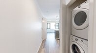 Located at 36 Traveler St in Boston's South End neighborhood (02118), this modern single-floor unit spans 300 square feet with a well-designed, efficient layout. The open-concept living and kitchen area features light hardwood-style flooring throughout, crisp white walls, and a kitchen island with a white quartz countertop and stainless steel sink. The kitchen is equipped with stainless steel appliances, including a refrigerator and microwave, complemented by sleek dark cabinetry and a subtle tile backsplash. Pendant lighting above the island adds a stylish touch, while recessed ceiling lights provide even illumination across the space. A separate bedroom area offers natural light through large windows with roller shades, providing views of the surrounding Boston cityscape. The hallway connects the living areas to the well-appointed bathroom, which features hexagonal gray tile flooring, a wood-toned vanity with a gray countertop and undermount sink, and a glass-enclosed walk-in shower with textured white tile walls. Chrome fixtures and overhead cabinetry complete the bathroom's contemporary design. Throughout the unit, clean lines, neutral tones, and modern finishes create a cohesive and functional living environment.