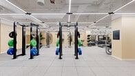Located at 705 W 9th St in Downtown Los Angeles, this expansive fitness facility spans a single floor and offers a comprehensive, well-equipped workout environment designed to accommodate a wide range of training styles. The open floor plan is finished with durable rubber sport tile throughout, providing both safety and functionality across all zones.

The free weights area is stocked with an extensive dumbbell rack, multiple adjustable benches, and wall-to-wall mirrors that amplify the sense of space and allow for form monitoring. A dedicated strength training section houses a row of Octagon power racks loaded with color-coded bumper plates, offering ample room for Olympic lifting and compound movements. A functional training zone features a heavy bag, cable attachments, and resistance tools, while a Matrix cable machine and cardio equipment serve users seeking versatile conditioning options.

The facility is illuminated by linear LED lighting suspended from an exposed ceiling, creating a bright and energizing atmosphere. Warm, neutral-toned wall panels add a refined aesthetic throughout. A glass-partitioned lounge area provides a relaxed retreat, furnished with mid-century style armchairs and round dining tables — ideal for recovery or informal gatherings. Built-in shelving and storage solutions are integrated throughout, keeping the space organized and accessible.