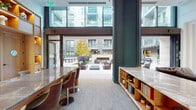 Located on the iconic Sunset Boulevard in the heart of Hollywood, this single-level lounge and amenity space at Hanover Hollywood seamlessly blends refined interior design with an expansive open-air courtyard. The interior lounge greets visitors with large-format tile flooring, warm wood built-in shelving displaying curated decorative objects, and a polished marble reception counter. A dedicated wet bar area features striking antique mirror wall panels, a mounted flat-screen display, and sleek wood cabinetry with under-counter refrigeration — ideal for social gatherings and events. Floor-to-ceiling glass walls and sliding doors dissolve the boundary between indoors and outdoors, flooding the interior with natural light and offering direct views into the central courtyard. The expansive outdoor space serves as the social hub of the property, featuring multiple seating arrangements including plush sectional sofas, lounge chairs, and a billiards table. An outdoor kitchen equipped with a stainless steel grill provides al fresco entertaining options, while an outdoor media wall with built-in screen, lush planted beds, mature trees in oversized planters, and string lights overhead create a vibrant yet relaxed atmosphere throughout.
