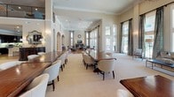 This elegant single-story community clubhouse is thoughtfully designed to offer a refined and welcoming environment for gathering and relaxation. The heart of the space is a grand, open hall with soaring ceilings, ornate chandelier lighting, and a mezzanine-level balcony that adds architectural drama. Arched doorways and crown molding throughout lend a classic, upscale character, while neutral tones and plush carpeting create a warm and inviting ambiance.

The main gathering area is generously proportioned, accommodating multiple large dining tables with upholstered chairs, alongside comfortable lounge seating arranged around a flat-screen entertainment center. Floor-to-ceiling windows and glass doors flood the space with natural light and offer direct views of the outdoor pool area, seamlessly blending indoor and outdoor living.

A dedicated lounge nook features an upright piano, plush seating, and dramatic patterned drapery framing oversized windows — an ideal retreat for quiet conversation or entertainment.

Two distinct kitchen areas serve the space: one with dark cabinetry, granite countertops, a stainless steel appliance suite, and hardwood-style flooring; the other a casual bar-style kitchen with a granite island, pendant lighting, a wine cooler, and a coffee station — perfect for hosting events of any scale.