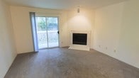 This single-floor residence offers a comfortable and well-appointed living space designed with both function and warmth in mind. The living area serves as the heart of the home, highlighted by a striking fireplace framed in a clean white marble surround — an elegant focal point that anchors the room. Soft, neutral-toned carpeting extends throughout, lending a warm and inviting atmosphere underfoot. The space is bathed in natural light courtesy of a large sliding glass door that opens directly onto a private outdoor deck. Beyond the deck, mature greenery and lush foliage create a serene, natural backdrop, providing both privacy and a refreshing connection to the outdoors. The walls are finished in a crisp, bright white, enhancing the sense of openness and allowing natural light to bounce freely throughout the interior. Overhead, a centrally placed light fixture provides additional illumination, complementing the abundant daylight. The overall ambiance is calm and neutral, offering a versatile canvas well-suited for a variety of interior styles. The seamless flow between the indoor living space and the outdoor deck makes this residence particularly appealing for those who value easy access to fresh air and nature.