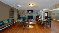 Located at 200 Branchwood Dr in Goose Creek, SC, this single-floor space offers an open, welcoming layout defined by rich hardwood flooring and impressively high ceilings with shiplap wood paneling that lend the interior a warm, rustic character. Natural light pours in through a large arched window above the entryway and a series of clerestory windows along the upper walls, creating a bright and airy atmosphere throughout the day. The main gathering area is expansive, comfortably accommodating multiple seating arrangements, including plush leather sofas and armchairs accented with teal throw pillows, arranged around a central coffee table. A dramatic floor-to-ceiling stone fireplace serves as the room's focal centerpiece, with a wall-mounted television above it, making it a natural hub for relaxation and socializing. Adjacent to the main seating area, a dining space features glass-top tables with cushioned chairs, ideal for shared meals or casual gatherings. A small beverage station with shelving and appliances is conveniently positioned nearby. The bold burgundy front door with leaded glass detailing makes a striking first impression at the entrance. Additional rooms branch off from the main living area, offering private or semi-private spaces. Ceiling fans and ample windows with blinds ensure comfort and light control throughout.