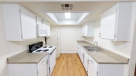 This single-floor space is designed with practicality and clean aesthetics in mind. The kitchen serves as a standout feature, laid out in a classic galley configuration that maximizes efficiency within its footprint. White shaker-style cabinetry lines both sides of the kitchen, offering generous storage space above and below the countertops. The countertops feature a neutral speckled finish that complements the crisp, white palette throughout. Appliances include a white electric range with coil burners, an over-the-range microwave, a full-size refrigerator, and a stainless steel double-basin sink with a modern faucet. A recessed tray ceiling with a built-in flush light fixture adds architectural interest overhead while providing bright, even illumination. Warm-toned hardwood-style flooring runs the length of the kitchen, adding a natural element that balances the otherwise cool, neutral tones of the space. The walls are finished in a soft, warm white that enhances the sense of openness and cleanliness. Overall, the space presents a tidy, move-in-ready environment that prioritizes functionality while maintaining a welcoming and well-lit atmosphere.