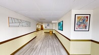 This single-floor building lobby and corridor offers a clean, functional, and welcoming environment designed to serve residents and visitors with efficiency and style. The space is finished with light gray wood-look flooring that runs the length of the corridor, complemented by two-tone walls featuring a warm yellow-cream upper section and a rich dark brown chair rail and wainscoting below, lending a structured yet inviting aesthetic throughout.

The drop-panel ceiling with recessed and flush-mounted lighting provides consistent, bright illumination across the entire space. A set of glass double-entry doors at the front floods the entryway with natural light and offers views of the surrounding outdoor area, creating an open and transparent point of arrival.

Moving through the corridor, curated artwork — including abstract geometric pieces and colorful framed prints — adorns the walls at regular intervals, adding character and visual interest. A dedicated mailroom area houses a large multi-unit mailbox panel, a decorative framed mirror, and a console table, providing residents with a convenient and organized mail retrieval point. An elevator is also accessible from within the corridor. A distinctive gold accent side table near the corridor's midpoint serves as a subtle decorative focal point. Overall, the space strikes a balance between practicality and polished design.