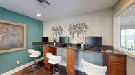 This single-floor space combines practical amenities with stylish design elements throughout its distinct areas. The business center is a standout feature, offering a long granite-topped workstation with warm wood cabinetry and multiple computer workstations. Modern white swivel chairs provide comfortable seating, while the teal accent wall adds a vibrant pop of color against neutral tones. Decorative metal tree wall art enhances the welcoming atmosphere, and recessed lighting ensures a well-lit working environment.

The dining area exudes an eclectic, artistic character centered around a striking round green marble pedestal table surrounded by woven rattan chairs, all set atop a natural fiber area rug over dark hardwood-style flooring. A large framed abstract artwork anchors the cream-colored wall, adding a gallery-like sophistication to the room. A sculptural oversized chalice-style centerpiece sits atop the table, serving as a bold decorative focal point.

Connectivity between rooms is achieved through distinctive architectural details, including a sliding barn door and a set of aged, rustic glass-paned doors, lending an industrial-chic aesthetic to the overall design. Recessed lighting throughout provides consistent, clean illumination, while the neutral wall palette allows the curated furnishings and art to take center stage.