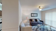 This 1-bedroom, 1-bathroom residence is thoughtfully designed to maximize comfort and functionality across its single-floor layout. The living room features soft carpet flooring, a ceiling-mounted light fixture, and a sliding glass door fitted with vertical blinds that allows natural light to fill the space. A defined dining area sits adjacent to the living room, separated by a charming spindle rail divider and illuminated by track lighting overhead, offering an intimate setting for meals. The galley-style kitchen is efficiently organized with dark wood cabinetry, white countertops, tile backsplash, and white appliances including a refrigerator, range, and dishwasher. Louvered bifold doors provide access to additional storage or utility space within the kitchen. The bedroom is a serene retreat with warm, neutral tones, plush carpet, and soft ambient lighting. The full bathroom features tile flooring, a vanity with cabinet storage, a toilet, and a tub-shower combination with a patterned curtain. Throughout the unit, neutral wall tones and classic wainscoting-style trim details create a cohesive, understated aesthetic that is both welcoming and easy to personalize.