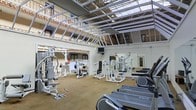 This fitness center is housed within a striking, high-ceilinged space characterized by an exposed steel roof structure and expansive skylights that flood the interior with natural light, creating an open and invigorating atmosphere. The facility is surrounded by upper-level balconies with warm-toned architectural detailing, adding a distinctive character to the workout environment. The main floor is generously sized, accommodating a comprehensive selection of strength training machines, including multi-function cable systems, weight benches, and plate-loaded equipment, all finished in a consistent white and grey palette. Cardio machines, including treadmills from True Fitness, are positioned toward the front of the space. Large mirrors line sections of the walls, enhancing the sense of spaciousness and allowing users to monitor their form. The flooring consists of a neutral-toned carpet, providing a practical and comfortable surface throughout. A wall-mounted television is also present, offering entertainment during workouts. The overall ambiance is bright, open, and well-equipped, making it a functional and visually appealing facility.