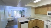 This single-floor residence is designed with an open-concept layout that seamlessly connects the kitchen and living areas, creating a cohesive and functional living environment. The kitchen is equipped with warm oak-toned cabinetry providing ample storage, a double-basin stainless steel sink, and a dishwasher integrated beneath the peninsula counter. The light-toned countertops extend across the peninsula, which serves as a natural divider between the kitchen and the adjoining living space. A skylight positioned above the kitchen enhances the area with additional natural illumination, complementing the recessed lighting in the ceiling. The textured ceiling adds a classic residential character throughout. The living area features carpeted flooring and is anchored by a large sliding glass door that opens to an outdoor patio, flooding the interior with abundant natural light and offering views of the surrounding greenery. A tall indoor plant near the patio doors adds a touch of organic warmth to the space. The overall ambiance is bright, open, and welcoming, with a practical flow between the kitchen and living zones suited for everyday living.