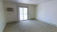 This 1-bedroom, 1-bathroom unit is laid out across a single floor, offering a functional and straightforward living arrangement. The living area is finished with wall-to-wall carpeting in a neutral tone, providing a soft and comfortable underfoot feel that complements the light-colored walls throughout. A sliding glass door serves as a prominent architectural feature, drawing in generous amounts of natural light and providing direct access to a private balcony with outdoor views of surrounding greenery. Sheer white curtains frame the door, softening the light while maintaining an airy feel. Baseboard heating runs along the perimeter of the room, offering an efficient and unobtrusive heating solution. The walls are finished in a clean, neutral palette that creates a calm and versatile backdrop, suitable for a variety of interior styles. The overall ambiance is understated and practical, with a focus on livability and simplicity. The single-floor layout ensures easy accessibility throughout the space, making it a straightforward and comfortable option for everyday living.