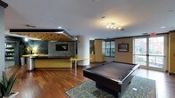 This single-floor recreational space is thoughtfully designed to offer a dynamic and social environment. The room is anchored by a full-size pool table set atop a decorative area rug, making it a natural focal point for entertainment and gathering. Rich hardwood flooring extends throughout, lending warmth and continuity to the space.

A standout feature is the bar and lounge area, distinguished by vibrant yellow cabinetry and countertops that create a bold visual contrast against the neutral walls. A dramatic textured stone accent wall frames a wall-mounted flat-screen television, adding a rustic yet refined element to the design. Overhead, a striking slatted wood ceiling detail adds depth and visual interest to the bar zone.

Structural columns are integrated seamlessly into the layout, contributing to the open yet defined spatial flow. Natural light filters in through multiple windows fitted with horizontal blinds, complementing the track lighting above that provides focused illumination throughout the room. Decorative artwork adorns the walls, adding a curated, finished feel.

The overall ambiance is modern and inviting, with a cohesive color palette of yellow, warm brown, and white that strikes a balance between energetic and comfortable—ideal for a community lounge or amenity space.