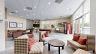 Located at 102 N Lemon Ave in Ontario, California, this single-floor community lounge offers a generously sized, multi-functional gathering space designed for both relaxation and recreation. The main area is anchored by a slate tile floor with decorative mosaic inlay borders, giving the space a polished, cohesive aesthetic throughout.

The lounge is divided into several distinct zones. A comfortable seating area features patterned armchairs and a sectional sofa arranged around a coffee table, complemented by a wall-mounted television and a built-in wood shelving unit displaying decorative accents. Adjacent to this is a game area centered around a full-sized pool table, positioned beside large floor-to-ceiling windows that flood the space with natural light and offer views of the surrounding outdoor greenery.

A well-equipped kitchenette serves the space, finished with rich dark wood cabinetry, granite countertops, a mosaic tile backsplash, stainless steel appliances, and pendant lighting. High-top dining tables and bar-height seating provide additional gathering spots near the windows.

The hallway connects to an elevator and staircase, with warm neutral walls, recessed lighting, and decorative wall-mounted wood shelving that adds a refined touch. Throughout the space, framed artwork and landscape photography contribute to an inviting, community-oriented ambiance.