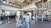The Peridot Palms Fitness Center in Saint Petersburg, FL is a thoughtfully designed two-story facility spanning 3,680 square feet across two floors — 1,970 sq ft on the ground level and 1,710 sq ft on the upper level. A dramatic steel spiral staircase serves as a central architectural focal point, connecting both floors with visual elegance beneath soaring double-height ceilings. The ground floor houses a dedicated functional training zone featuring resistance ropes, a boxing dummy, suspension trainers, and cable machines, all set against a striking exposed brick accent wall. A mirrored free weights area with a full dumbbell rack adjoins this space. A separate spin studio on this level accommodates multiple cycling bikes alongside medicine ball and stability ball storage, with large windows allowing natural light to fill the room. The upper level features a well-equipped cardio floor lined with treadmills, ellipticals, and stair climbers from brands including Precor and Cybex, bathed in natural light from expansive floor-to-ceiling windows offering outdoor views. A welcoming lounge area with seating, wood-look flooring, and a blue accent rug bridges zones on the upper level, alongside built-in storage lockers. The facility's modern aesthetic is elevated throughout by recessed lighting, ceiling fans, and clean neutral tones.