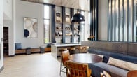 This single-level amenity space delivers a sophisticated blend of industrial and contemporary design. The centerpiece is a grand lounge area with soaring ceilings clad in warm wood paneling, framed by bold dark vertical steel columns and floor-to-ceiling windows that flood the interior with natural light and offer views of a landscaped outdoor courtyard. A sleek bar island with bar stool seating sits beneath dramatic black dome pendant lights, flanked by built-in open shelving displaying curated décor. A welcoming seating lounge features plush sofas, a wood-slab coffee table, and a floor-to-ceiling vertical slat partition that elegantly divides zones. A stone-clad fireplace with stacked log storage and a wall-mounted television anchors a cozy lounge area, with large sliding glass doors opening directly to an outdoor patio. A dedicated game room offers a pool table, shuffleboard, and wall-mounted TV, adjacent to a glass-walled fitness center. A secondary lounge and dining nook feature a blue shiplap accent wall adorned with eclectic artwork. Light wood-look tile flooring flows throughout, completing the cohesive, upscale aesthetic of this well-appointed communal space.