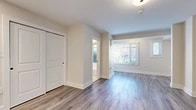 This single-floor space is thoughtfully designed with a clean, modern aesthetic that carries consistently throughout every room. The living area is bathed in natural light streaming through large bay windows fitted with white horizontal blinds, offering views of mature trees and greenery outside. Warm-toned, light grey hardwood-style flooring extends seamlessly across the entire unit, lending a cohesive and polished feel underfoot.

The open layout connects the living space to a well-appointed kitchen featuring white shaker-style cabinetry, a white subway tile backsplash, and stainless steel appliances including a gas range and a full-sized refrigerator. Recessed lighting illuminates the kitchen efficiently, while a sleek black-fixtures sink adds a contemporary accent. A full-panel closet door adjacent to the kitchen provides convenient additional storage.

The living area is further defined by architectural column details and a neatly recessed wall niche, adding subtle character to the otherwise clean lines of the space. Neutral warm-white walls throughout create a calm, versatile backdrop.

The bathroom is finished with classic black-and-white mosaic floor tile, a white shaker vanity with black hardware, a medicine cabinet mirror, and a full bathtub, delivering both timeless style and practical functionality. The overall ambiance of this space is fresh, bright, and move-in ready.