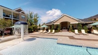 This single-story community clubhouse offers a well-rounded amenity experience, blending indoor functionality with an inviting outdoor retreat. The exterior is highlighted by a classic architectural design featuring a gabled roofline, stone accents, and a covered entryway framed by lush climbing greenery and manicured landscaping. A generously sized outdoor pool serves as the focal point, complete with a bubbling water feature and a wide, open sundeck. Lounge chairs and a shaded umbrella area provide ample space for relaxation under open skies, making it an ideal setting for outdoor leisure. Inside, the business center is thoughtfully designed for productivity and convenience. Accessed through glass-paneled doors, the room features built-in granite-top workstations arranged in an L-shaped configuration, accommodating multiple workstations with desktop computers. A dedicated printer station ensures residents have full office capabilities at their disposal. Crown molding and neutral wall tones give the space a clean, polished look, while typewriter-themed artwork adds a touch of character. Interior windows connect the business center to the adjacent common area, maintaining an open and connected feel throughout the clubhouse.