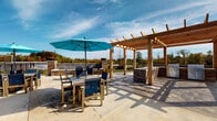 Located at 12207 Avoca Rd in Louisville, Kentucky, this single-level outdoor amenity space is thoughtfully designed to support both relaxed gatherings and active entertaining. The area is divided into distinct functional zones that flow seamlessly across a broad concrete patio.

A covered lounge area anchors one portion of the space, featuring a large wraparound sectional sofa with plush gray cushions accented by teal throw pillows. Ceiling fans overhead provide comfort during warmer months, and large black-framed windows along the building facade allow views between the interior and exterior spaces. Brick and white lap siding detailing on the building exterior adds a classic, polished aesthetic.

Extending beyond the covered section, a substantial wood pergola defines the open-air entertaining zone. Beneath and around it, an outdoor kitchen with stone accents and multiple stainless steel grills serves as a focal point for outdoor dining. Slatted dining tables with navy blue chairs, including an umbrella-shaded set, provide ample seating for guests.

The patio opens to sweeping views of mature trees with autumn foliage, and the surrounding landscape provides a natural backdrop. A gated pool area and additional community buildings are visible in the distance, reinforcing the scale and connectivity of this well-appointed outdoor community space.
