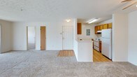 The Raven floor plan is a single-story, two-bedroom, one-bathroom unit designed with a practical and comfortable layout. The open living area is finished with neutral-toned carpeting and features a ceiling fan, providing a versatile space for both relaxing and entertaining. Visible from the living area, the kitchen is positioned adjacent to the entry and features warm wood cabinetry, white appliances including an electric range and refrigerator, light hardwood-style flooring, and ample counter space along a galley-style layout. A pass-through counter connects the kitchen to the living space, and an electrical panel is conveniently located within the kitchen.

Both bedrooms are carpeted and receive natural light through windows with blinds. The primary bedroom includes a ceiling fan and access to a walk-in closet, offering generous storage. The second bedroom is similarly bright and well-proportioned.

The full bathroom is equipped with a tub-shower combination, a vanity with wooden cabinetry, and tile flooring. A notable feature of this unit is the in-unit stacked washer and dryer, housed in a dedicated closet within or adjacent to the bathroom, adding everyday convenience. Throughout, textured ceilings and neutral wall tones contribute to a clean, move-in-ready aesthetic.