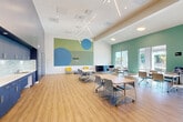 Located at 2144 Sheraton Dr in Santa Clara, California, this single-floor space is designed as a versatile, open-plan activity or community room that balances functionality with a welcoming aesthetic. The interior is defined by elevated ceilings that lend a sense of openness, complemented by warm wood-look flooring throughout. A striking large-scale mural featuring bold circles in shades of blue and green serves as a focal point on one wall, adding visual energy to the space.

Lighting is a standout feature, with a geometric linear pendant fixture suspended from the ceiling alongside recessed lighting, ensuring thorough, even illumination. The room accommodates multiple flexible tables on casters paired with mobile chairs, supporting a variety of seating configurations for group activities or meetings. Additional seating areas include a round table set near the large sliding glass doors, which open directly to an outdoor patio area and allow abundant natural light and exterior views to flow in.

A built-in kitchenette along one wall features bold navy-blue cabinetry, white countertops, a stainless steel sink, a glossy tile backsplash, and a full-size refrigerator, making the space suitable for events and gatherings. Storage lockers and a display unit round out the room's practical amenities, while the teal and white color palette throughout reinforces the space's modern, inviting character.