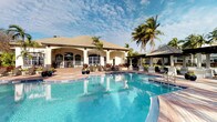 This single-story clubhouse facility presents a welcoming, resort-inspired environment centered around a generously sized outdoor swimming pool with crystal-clear turquoise water. The building itself showcases a Mediterranean-influenced architectural style, characterized by a warm beige stucco exterior, a hip roof with earthy-toned tiles, and a series of arched openings along the front façade that provide a graceful, open-air transition between the interior and the surrounding pool deck.

The expansive pool deck is finished with decorative pavers and is furnished with outdoor seating, creating comfortable areas for lounging and socializing. A separate covered pergola structure to the side of the main building offers a shaded retreat, ideal for gatherings out of direct sunlight. Tall, swaying palm trees and tropical plantings frame the space, enhancing the lush, vacation-like ambiance.

The interior of the clubhouse, visible through the arched openings, appears bright and airy, benefiting from natural light that filters in through large windows and the open colonnade. The overall setting conveys a relaxed yet polished atmosphere well-suited for community recreation and events.