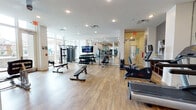 This modern fitness center offers a spacious and thoughtfully designed workout environment spread across a single floor. The space is finished with warm hardwood flooring throughout, white walls, and recessed lighting that creates a clean, bright atmosphere. Large floor-to-ceiling windows along one side of the facility flood the interior with natural light and provide views to the outdoors, contributing to an open and energizing ambiance. Full-length mirror walls amplify the sense of space and allow users to monitor their form during exercise. The cardio zone is well-appointed with multiple treadmills, elliptical machines, and stationary bikes, including Life Fitness and Matrix branded equipment. A dedicated strength training area features a full rack of dumbbells, adjustable weight benches, and a variety of cable and resistance machines, including Life Fitness multi-function units. A flat-screen television is mounted centrally, visible from multiple areas of the gym. Structural columns are integrated cleanly into the layout, preserving an unobstructed flow throughout the space. A glass-panel door provides a secondary access point, while designated areas near the entrance offer informational signage and storage. The overall design balances functionality with a contemporary aesthetic, making it a comprehensive and inviting facility for a full range of fitness activities.