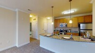 This single-floor residence is designed with a functional, open-concept layout that seamlessly connects the kitchen and living areas. The kitchen is a standout feature, showcasing warm honey-toned cabinetry, granite countertops, and a full suite of stainless steel appliances including a range, microwave, and refrigerator. A curved breakfast bar with pendant lighting overhead serves as both a dining and entertaining focal point, while tile flooring grounds the cooking space with a clean, durable finish. Crown molding adds a refined architectural detail throughout the main areas. The adjoining living space features plush wall-to-wall carpeting, creating a comfortable transition from the hard-surface kitchen. The bedroom is bright and welcoming, with two windows dressed in white blinds that allow ample natural light to fill the room, paired with the same soft carpeting underfoot. The bathroom is well-equipped with tile flooring, a tub-shower combination featuring full tile surround, a single-sink vanity with matching wood cabinetry, and a large mirror. Additional doors visible from the main living area suggest convenient closet or utility access. Overall, the space presents a cohesive, practical design with quality finishes throughout.