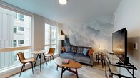This single-floor residence showcases a thoughtfully designed open-concept layout that seamlessly connects the living, dining, and kitchen areas. The living room is defined by a dramatic floor-to-ceiling watercolor mountain mural that serves as the focal point of the space, complementing the tufted gray sofa, mid-century modern coffee table, and warm accent furnishings. Large floor-to-ceiling windows flood the living and dining area with natural light, offering views of the surrounding urban environment. A compact dining nook with a round wood table and leather chairs sits adjacent to the windows, making efficient use of the space.

The kitchen features flat-front cabinetry extending to the ceiling in a warm off-white tone, paired with a subway tile backsplash and a full suite of stainless steel appliances, including a refrigerator, electric range, microwave, and dishwasher. Light wood-look flooring runs continuously throughout the unit, creating a cohesive and airy aesthetic.

The bedroom is a calm, neutral retreat with soft gray walls, wood nightstands, and warm ambient lighting. The bathroom offers a clean, modern design with an extended single-sink vanity topped in white, an illuminated mirror, glass-enclosed shower, and the same light wood flooring carried throughout the unit.