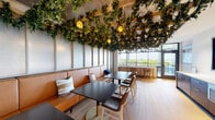 This single-floor lounge and dining space makes an immediate impression with its most striking architectural feature: a lush, sprawling canopy of cascading greenery suspended from a wood-lattice ceiling structure. Interspersed throughout the botanical installation are warm amber pendant lights that cast an inviting, garden-like glow across the entire room.

The seating area is anchored by continuous cognac leather banquette seating running along the perimeter walls, complemented by dark-topped dining tables and contemporary wood-framed chairs with cushioned seats. Textured wall paneling in a soft neutral tone adds depth and warmth to the surrounding walls, while light-toned wood-look tile flooring grounds the space with a clean, modern aesthetic.

Floor-to-ceiling windows along one wall flood the interior with abundant natural light and provide expansive treetop views of the surrounding landscape, creating a seamless connection between the lush interior greenery and the natural environment outside. A glass door leads to an adjacent outdoor area, further extending the indoor-outdoor feel.

A built-in kitchenette along one wall features dark wood cabinetry, a white countertop with sink, a microwave, and a wall-mounted flat-screen television. Curated artwork, including a vibrant large-scale painting, adds cultural character to the space. Together, these elements create a sophisticated, biophilic retreat ideal for gathering and dining.