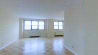 This single-floor space offers a clean, functional layout with a welcoming and airy atmosphere. The main living area is generously proportioned, characterized by a distinctive light-toned checkered tile floor that adds a classic charm to the room. Crisp white ceilings and neutral cream-colored walls create a bright, open feel, while multiple windows fitted with horizontal blinds allow an abundance of natural light to fill the space. Beneath each window, built-in radiator units provide efficient heating, reflecting the practical and well-maintained nature of the space. The open-plan design of the living area allows for flexible furniture arrangements and offers a sense of spaciousness. The smooth white ceiling and clean baseboards contribute to a tidy, well-kept aesthetic throughout. The layout and architectural details suggest a straightforward, classic design sensibility, making the space versatile and adaptable for a variety of uses. Overall, this space combines practical features with a bright, inviting ambiance, suitable for comfortable everyday living.