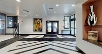 This single-floor commercial lobby space makes a striking first impression with its bold black-and-white geometric tile flooring, which creates dynamic visual pathways throughout the open layout. The main entrance area is anchored by a set of glass double doors and accented by a large, colorful abstract painting that adds artistic character to the bright white walls.

The space is thoughtfully divided into distinct functional zones. A lounge seating area features a curated mix of orange velvet armchairs and patterned accent chairs arranged around sleek gold-legged side tables, all set against a textured white feature wall adorned with stacked spherical brass wall sconces. A second seating nook offers a plush purple sofa, a glass-top coffee table, and an area rug, positioned alongside floor-to-ceiling windows dressed in warm-toned sheer vertical drapes that invite generous natural light and outdoor views.

Adjacent to the lounge area, a dedicated workstation zone provides a row of individual desk spaces equipped with ergonomic chairs, ideal for focused tasks or informal meetings. The space is finished with a suspended tile ceiling, recessed lighting, and clean white walls with contrasting dark baseboards. Restroom access is also available within the layout. The overall ambiance is modern, polished, and welcoming, balancing professional functionality with design-forward aesthetics.