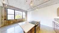 This single-floor urban loft blends industrial character with contemporary finishes to create a striking and functional living space. The exposed concrete ceiling and polished concrete floors serve as defining architectural elements, lending the space an authentic industrial aesthetic that is both raw and refined. Original brick detailing along the walls adds warmth and texture, paying homage to the building's heritage while complementing the modern upgrades throughout.

The kitchen is a standout feature, centered around a generous island topped with elegant white quartz countertops accented by subtle veining. A sleek black electric range with stainless steel accents is integrated into the island, making it as practical as it is visually impressive. Above, a pair of geometric brass-framed pendant lights cast a warm, ambient glow that enhances the space's modern industrial character.

Large black-framed windows allow generous natural light to pour in, illuminating the open-plan layout and offering views of surrounding greenery and the urban streetscape beyond. The open floor plan creates a seamless flow throughout, providing a versatile canvas for a variety of living arrangements. The combination of soaring ceilings, natural light, and thoughtful material choices gives this loft an airy, spacious feel that is both inviting and sophisticated.
