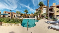 Located at 8330 N 19th Ave in Phoenix, Arizona, this outdoor amenity space serves as the central gathering hub of a multi-building residential community. The centerpiece is a generously sized freeform swimming pool with crystal-clear turquoise water, accented by a natural rock waterfall feature that adds a resort-like ambiance. A separate spa sits adjacent to the main pool, connected by a smooth concrete pool deck that provides ample space for lounge seating and relaxation under the Arizona sun. Mature fan palms and desert-adapted landscaping are strategically placed throughout the space, offering both shade and visual appeal while reinforcing the Southwestern aesthetic. A dedicated poolside structure with a tile roof serves as a cabana or clubhouse, featuring louvered panels and outdoor seating, ideal for gatherings or shade. The surrounding community buildings display a warm stucco exterior with Spanish-influenced rooflines and private balconies overlooking the amenity area. A gated children's play zone with a turf surface and playground equipment is nestled within the complex, enclosed by decorative iron fencing for safety. Covered pergola entryways and desert rock ground cover complete the thoughtfully designed outdoor environment, making this a well-appointed communal space built for year-round enjoyment in the Phoenix climate.