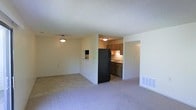 This single-floor residence offers a straightforward, functional layout designed for comfortable everyday living. The main living area is generously sized, featuring wall-to-wall carpet flooring that adds warmth underfoot. A flush-mount ceiling light fixture provides overhead illumination, while a small built-in wall niche adds a subtle architectural detail. The space is finished with neutral-toned walls and a textured ceiling, contributing to a clean and understated aesthetic. A floor vent ensures efficient climate control throughout. The living area flows naturally toward the kitchen, which is partially separated by a half-wall partition. The kitchen is outfitted with medium-toned wood cabinetry, a dark-finish refrigerator, and a sink set beneath the cabinetry. Tile flooring in the kitchen contrasts with the carpeted living space, clearly delineating the two zones. Recessed and flush ceiling lighting in the kitchen ensures the area is well-lit for meal preparation. The overall layout is efficient and practical, making good use of the available space across a single level. The neutral color palette throughout creates a cohesive and move-in-ready environment.