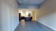 This single-floor space showcases a thoughtfully designed open-concept layout that seamlessly connects the living area to the kitchen. The kitchen serves as a focal point, featuring a combination of dark espresso lower cabinetry and crisp white upper cabinets, creating a striking two-tone contrast. A movable dark island anchors the center of the kitchen, offering additional workspace and flexibility. Stainless steel appliances, including a refrigerator, microwave, and range, lend a sleek, contemporary feel. Track lighting mounted beneath the upper cabinets illuminates the workspace effectively.

One of the most notable architectural features is the soaring ceiling height, which is accentuated by exposed piping running overhead — a nod to industrial-style design. This elevated ceiling contributes to an airy, open atmosphere throughout the space. The flooring consists of durable wood-look planks in a cool gray tone, running consistently through both the living and kitchen areas to create a cohesive flow. Neutral gray walls complement the overall modern palette. The open layout between the kitchen and living space makes this unit highly functional and well-suited for everyday living.