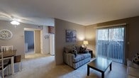 The Rosebud unit is a well-appointed 1-bedroom, 1-bathroom residence spread across a single floor, offering a practical and comfortable layout. The living area serves as the heart of the home, featuring warm neutral-toned walls and plush carpeting that creates an inviting atmosphere. A large sliding glass door dressed with vertical blinds allows natural light to fill the space while providing access to the outdoors. The open sightlines connect the living room to the adjacent areas, enhancing the sense of space within the floor plan. A partial wall divides the living space from the utility and kitchen areas, where a full-sized refrigerator is visible, indicating a well-equipped kitchen setup. A doorway leads to additional rooms, including the bedroom and bathroom, maintaining a smooth flow throughout the unit. The textured ceiling adds a classic touch to the interior, complementing the understated décor. Overall, the unit presents a clean, functional, and comfortable living environment suited for individuals or couples seeking a manageable, single-level home with key amenities close at hand.