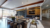 This thoughtfully designed single-floor amenity space seamlessly blends modern industrial aesthetics with warm, rustic elements to create an inviting and multifunctional environment. Exposed wood ceiling beams run throughout, complementing the rich wood wall paneling and large-format gray tile flooring that anchor the design palette.

The spacious lounge area serves as the centerpiece, featuring a sleek electric fireplace framed by a wood-panel surround and a wall-mounted flat-screen TV above. A dramatic floor-to-ceiling dark shelving unit adds depth and visual interest, while a statement chandelier with an orb silhouette illuminates generous seating arrangements ideal for gathering and relaxation.

Adjacent to the lounge, a well-appointed wet bar area showcases dark granite countertops, light wood cabinetry, a subway-style brick tile backsplash, open shelving, and stainless steel appliances. A tufted burnt-orange upholstered accent wall adds a bold pop of color alongside casual dining seating, completing this entertaining-ready nook.

The lobby entry is airy and open, with large windows flooding the space with natural light. Sputnik-style chandeliers and pendant lights create a striking visual display overhead. Workstation desks with black metal-framed glass partitions provide functional areas for residents or staff. A secondary seating vignette with brick accent walls and a wall-mounted TV rounds out the welcoming atmosphere of this refined communal space.