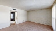 This single-floor, 2-bedroom, 1-bathroom residence offers a practical and well-organized layout. A defining architectural feature throughout the home is the painted white stacked-brick accent wall, which appears in multiple rooms, lending a consistent and distinctive character to the interior. The living room is a generously sized carpeted space with a textured ceiling, ceiling-mounted HVAC venting, and a sliding glass door that allows natural light to filter in through vertical blinds, offering a view to an outdoor area. The primary bedroom is a comfortable, well-proportioned room with the same painted brick wall, carpet flooring, a ceiling fan with light fixture, and a window with blinds. Both bedrooms feature sliding closets with built-in shelving and dark wood-toned doors, providing ample storage. The kitchen runs along a galley-style layout with warm wood cabinetry, laminate countertops, a double sink, and wood-look vinyl flooring. A window above the countertop brings in natural light. White appliances include a refrigerator, range, and dishwasher. The bathroom is fitted with a tub-shower combination, white tile surround, chrome fixtures, and a frosted window for privacy. A hallway connects the main living areas to the bedrooms and bathroom, completing this compact yet functional floor plan.