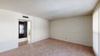 This single-floor, 2-bedroom, 1-bathroom residence offers a practical and well-organized layout. A defining architectural feature throughout the home is the painted white stacked-brick accent wall, which appears in multiple rooms, lending a consistent and distinctive character to the interior. The living room is a generously sized carpeted space with a textured ceiling, ceiling-mounted HVAC venting, and a sliding glass door that allows natural light to filter in through vertical blinds, offering a view to an outdoor area. The primary bedroom is a comfortable, well-proportioned room with the same painted brick wall, carpet flooring, a ceiling fan with light fixture, and a window with blinds. Both bedrooms feature sliding closets with built-in shelving and dark wood-toned doors, providing ample storage. The kitchen runs along a galley-style layout with warm wood cabinetry, laminate countertops, a double sink, and wood-look vinyl flooring. A window above the countertop brings in natural light. White appliances include a refrigerator, range, and dishwasher. The bathroom is fitted with a tub-shower combination, white tile surround, chrome fixtures, and a frosted window for privacy. A hallway connects the main living areas to the bedrooms and bathroom, completing this compact yet functional floor plan.