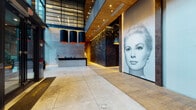 This expansive lobby is defined by its dramatic architectural design and carefully curated artistic elements. Soaring double-height glass curtain walls flood the space with natural light, framing views of the surrounding streetscape while reinforcing a sense of openness and transparency. Large-format concrete tile flooring grounds the space with an industrial yet refined aesthetic.

A monumental three-dimensional sculptural wall anchors the reception area, composed of layered dark metallic fragments arranged in an intricate, textured relief. Overhead, a warm wood-paneled ceiling contrasts beautifully with the cool concrete and steel palette below. Bold cylindrical pendant lights with vivid orange interiors provide warm accent illumination throughout.

A large-scale black-and-white portrait artwork commands attention along one wall, becoming a defining visual focal point of the lobby. A secondary seating area features a lounge sofa positioned beside a fireplace, accompanied by a dramatic steel staircase and full-height glazing. Adjacent to the main hall, a dedicated mail room is appointed with crystal chandelier lighting, wooden sorting consoles, and an expressive abstract painting. Illuminated display niches with orange-toned backlighting add refined decorative detail throughout.