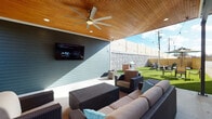 Located at 180 N Martin Luther King Blvd in Lexington, Kentucky, this thoughtfully designed outdoor space combines modern architecture with functional entertaining areas. The building exterior presents a striking combination of red brick and blue-grey horizontal siding, accented by manicured planters and evergreen shrubs that frame the entryway along a clean concrete walkway.

The centerpiece of the space is a generously sized covered patio featuring a warm wood-paneled ceiling with recessed lighting and a large ceiling fan, creating a comfortable environment regardless of weather. The lounge area is furnished with wicker seating, including a sectional sofa and armchairs arranged around a coffee table, all oriented toward a wall-mounted flat-screen television — ideal for outdoor gatherings and entertainment.

Adjacent to the covered lounge, an open-air lawn area showcases lush artificial turf, bordered by a built-in brick grill station equipped with a stainless steel grill. String lights are strung above, adding ambiance for evening use. A stone-textured perimeter wall with horizontal wood privacy fencing encloses the space, providing a sense of seclusion while maintaining an open, airy feel. A separate dining area with bar-height seating rounds out the versatile outdoor layout.