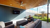 Located at 180 N Martin Luther King Blvd in Lexington, Kentucky, this thoughtfully designed outdoor space combines modern architecture with functional entertaining areas. The building exterior presents a striking combination of red brick and blue-grey horizontal siding, accented by manicured planters and evergreen shrubs that frame the entryway along a clean concrete walkway.

The centerpiece of the space is a generously sized covered patio featuring a warm wood-paneled ceiling with recessed lighting and a large ceiling fan, creating a comfortable environment regardless of weather. The lounge area is furnished with wicker seating, including a sectional sofa and armchairs arranged around a coffee table, all oriented toward a wall-mounted flat-screen television — ideal for outdoor gatherings and entertainment.

Adjacent to the covered lounge, an open-air lawn area showcases lush artificial turf, bordered by a built-in brick grill station equipped with a stainless steel grill. String lights are strung above, adding ambiance for evening use. A stone-textured perimeter wall with horizontal wood privacy fencing encloses the space, providing a sense of seclusion while maintaining an open, airy feel. A separate dining area with bar-height seating rounds out the versatile outdoor layout.