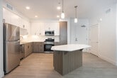 Located at 18831 Von Karman Ave in Irvine, CA, this well-appointed 2-bedroom, 2-bathroom unit offers a thoughtfully designed single-floor layout. The open-concept kitchen and living area serves as the heart of the home, featuring white upper cabinetry paired with warm wood-tone lower cabinets, a white quartz countertop, and a distinctive geometric kitchen island with pendant lighting overhead. Stainless steel appliances — including a refrigerator, gas range, microwave, and dishwasher — complete the kitchen. Light wood-style flooring flows throughout the main living areas, complementing the bright, clean aesthetic. The living room benefits from natural light and direct access to a private balcony through a glass door. Both bedrooms are carpeted and include ceiling fans and walk-in closets. The primary bedroom is a comfortable, well-proportioned retreat. Each bathroom is finished with wood-tone vanity cabinetry, white quartz countertops, large mirrors, and a full shower-tub combination. The unit maintains a consistent modern design language throughout, with clean white walls, recessed lighting, and a neutral palette that creates a bright and airy ambiance across all rooms.