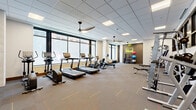 Located at 260 Portland Ave in downtown Minneapolis, this fitness center offers a well-appointed, single-floor space designed to support a comprehensive workout experience. The open layout provides generous room to move, accommodating a wide variety of equipment without feeling crowded.

The cardio zone lines the expansive floor-to-ceiling windows along one wall, housing multiple treadmills, elliptical machines, and a rowing machine. The natural light flooding in through these large windows creates an energizing atmosphere while offering views of the surrounding urban environment. Ceiling fans are positioned throughout to maintain comfortable airflow.

The strength training area features a cable machine, multiple weight racks with dumbbells, and a barbell station, providing ample options for resistance training. A dedicated functional fitness corner includes kettlebells, stability balls, and open floor space, accented by an exposed brick wall bearing a neon "Stay Hydrated" sign, adding a modern, motivating touch.

The entrance area is framed by sleek black-framed glass doors and walls, alongside a bold abstract mural that adds an artistic flair to the space. Patterned carpet tiles with subtle accent lines cover the floor throughout, while a suspended tile ceiling with recessed and panel lighting ensures the space is well-lit at all hours. The overall design balances contemporary aesthetics with practical fitness functionality.