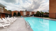 Aura is a well-appointed residential community offering single-floor living within a multi-building complex. The property is designed with both comfort and functionality in mind, providing residents with a range of on-site amenities and practical interior features.

At the heart of the community's outdoor space is a generously sized swimming pool, framed by a wide concrete deck equipped with lounge chairs and colorful seating. The pool area offers open views of the surrounding brick-exterior buildings, mature trees, and landscaped grounds, creating a welcoming atmosphere for outdoor relaxation.

Inside, the community includes a dedicated office or business center, accessible via a glass-paned door. This space is furnished with a workstation, computer, and wall art, providing residents with a clean, functional environment for work or administrative needs. The interior hallways feature neutral gray walls and hard-surface flooring, contributing to a modern and cohesive aesthetic throughout the common areas.

Utility spaces are also thoughtfully integrated, with utility closets housing essential mechanical equipment. Overall, Aura combines practical residential infrastructure with community-focused amenities, making it a functional and comfortable place to call home.