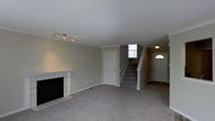This two-floor home presents a welcoming and functional layout designed with practical living in mind. The main level living room is finished with wall-to-wall carpet in a neutral grey tone, creating a comfortable and cohesive feel throughout the space. A tiled fireplace with a white mantel serves as a natural focal point along one wall, adding both character and warmth to the room. Track lighting mounted on the ceiling provides adjustable illumination, complementing the textured ceiling finish. Neutral-toned walls with white trim and baseboards give the space a clean, understated aesthetic. A central staircase with carpeted treads and a white newel post connects the main level to the upper floor, positioned alongside a window that allows natural light to filter into the stairwell. Adjacent to the staircase, the front entryway features a solid door with a decorative arched window above, enhancing natural light at the entrance. A secondary interior door on the main level provides access to an additional room or storage area. Overall, the space offers a straightforward, move-in-ready environment with classic residential finishes throughout.