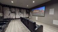 This private screening room is designed to deliver an immersive cinematic experience, combining thoughtful thematic design with high-quality audio-visual technology. The focal point of the space is a large wall-mounted display screen positioned on a neutral gray accent wall, flanked by flush-mounted in-wall speakers that form part of a comprehensive surround sound system. Additional speakers are integrated throughout the room, ensuring balanced audio coverage from every seat.

Seating is arranged in two tiered rows of black leather reclining chairs, each equipped with built-in cup holders, providing both comfort and a classic movie theater feel. The layout allows for clear sightlines to the screen from all positions.

The most distinctive design element is the full-scale film countdown mural that spans the rear and side walls, rendered in a vintage black-and-white aesthetic with film strip borders framing oversized countdown numerals. Wall-mounted sconce lighting accents the mural and contributes to the room's atmospheric ambiance. A bold red entry door with a porthole-style window serves as a striking contrast to the otherwise dark, moody interior.

The floor is finished with a textured, gray-toned carpet that complements the overall color palette. Recessed ceiling lighting provides practical illumination, completing this well-appointed, cinema-inspired space.